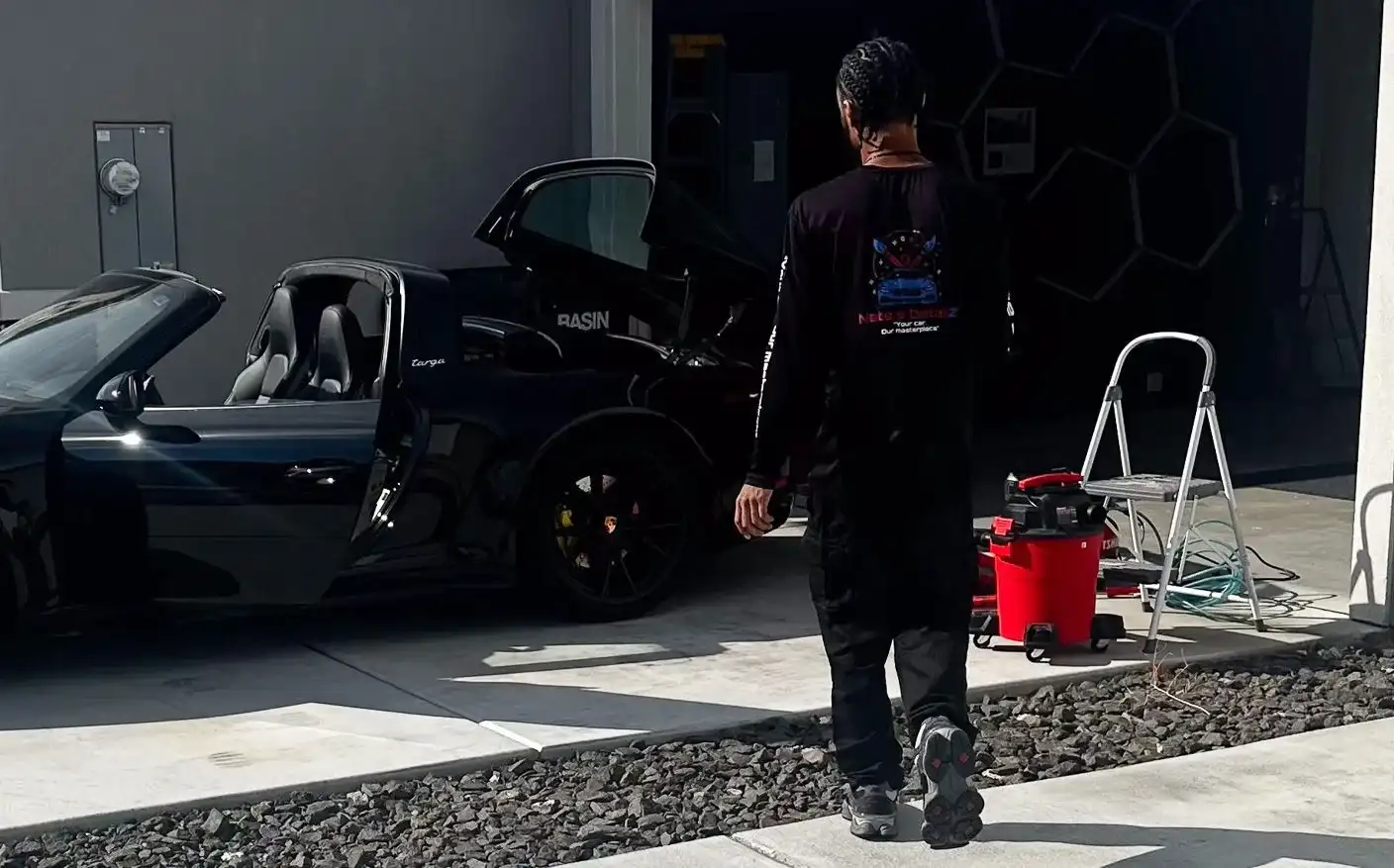 Nate, in his Nate’s Detailz shirt, walking past his mobile setup beside a black Porsche 911 with its doors open