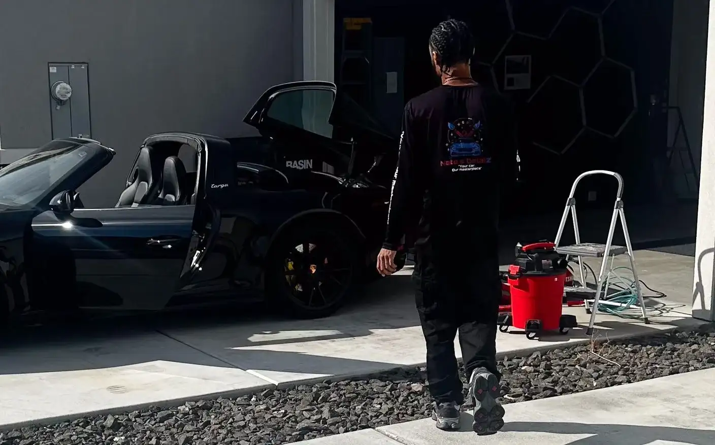 Nate, in his Nate’s Detailz shirt, walking past his mobile setup beside a black Porsche 911 with its doors open